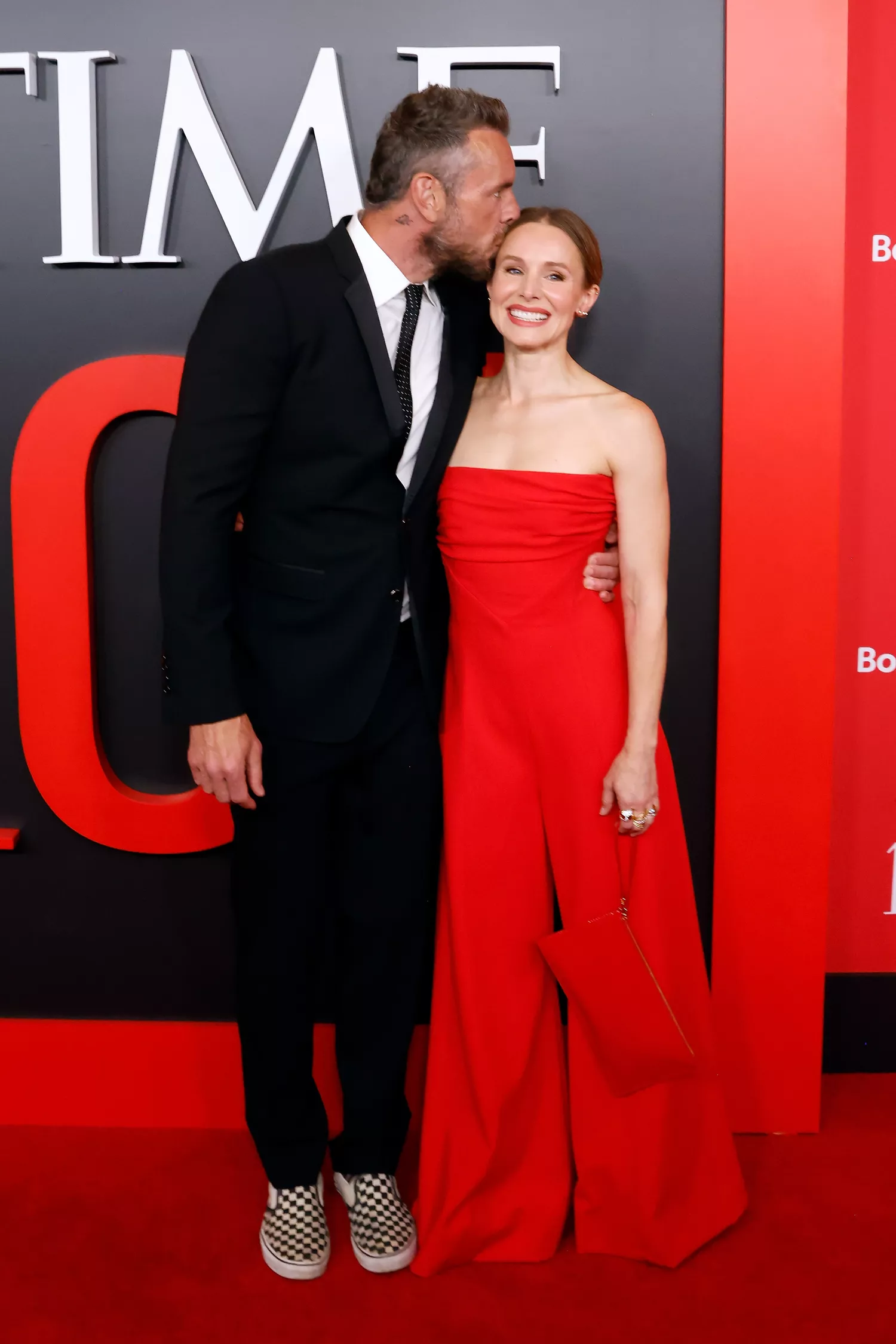 Kristen Bell and Dax Shepard attend the TIME100 Gala red carpet, sharing a sweet moment. Kristen Bell wears a red jumpsuit while Dax Shepard dons a classic suit.