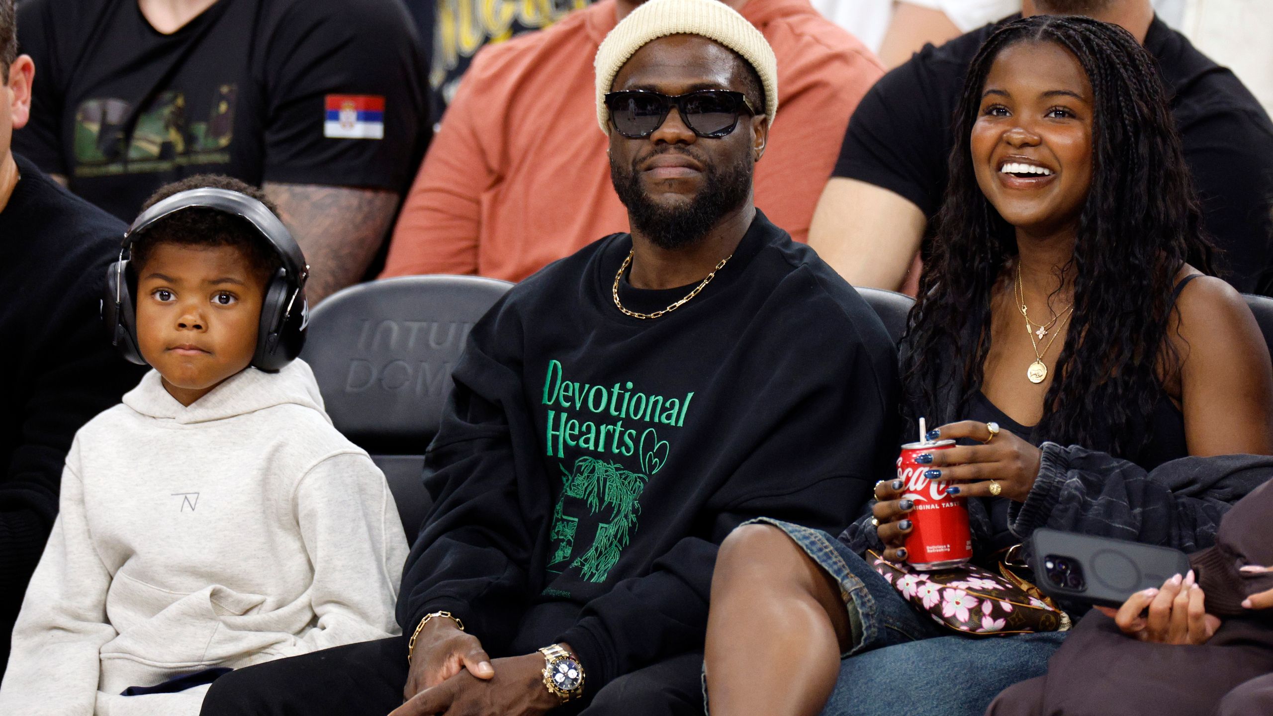 Kevin Hart smiling courtside at a basketball game, wearing a luxury Rolex watch